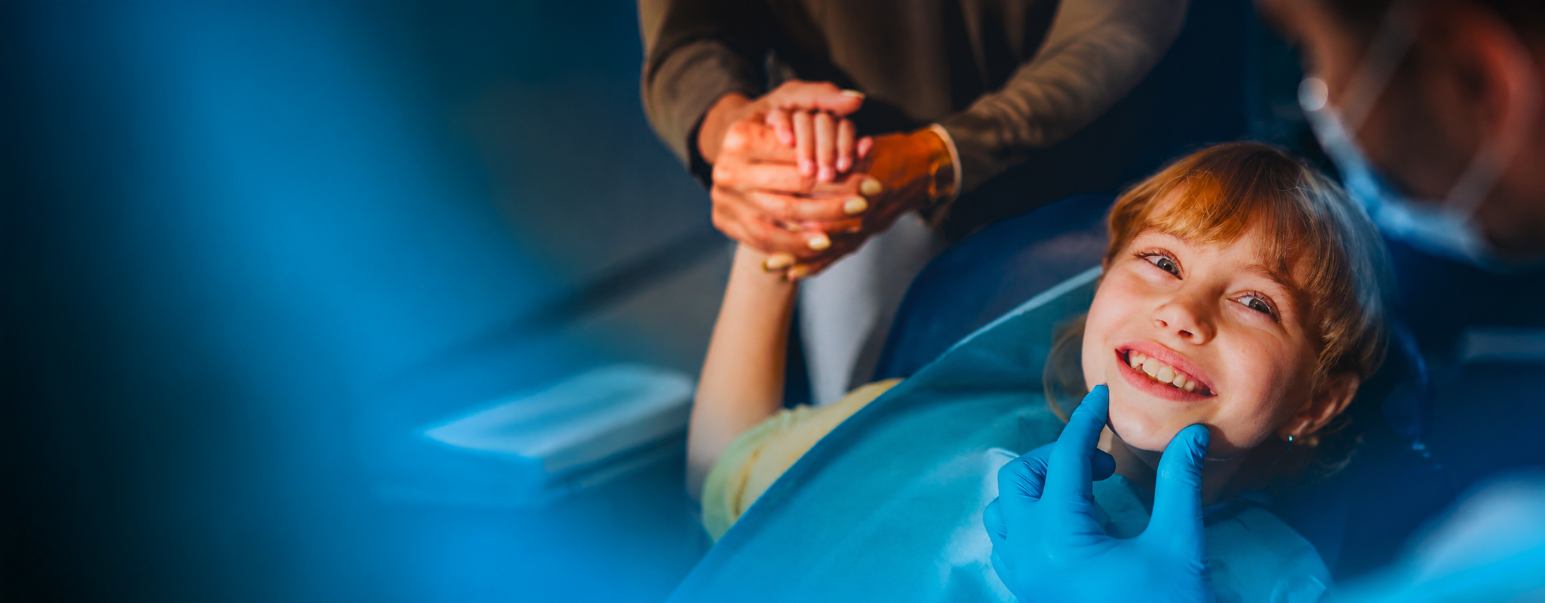 A young girl smiling in the dental chair, her parent holding her hand, in the deep blue glow of the ACTEON key visual